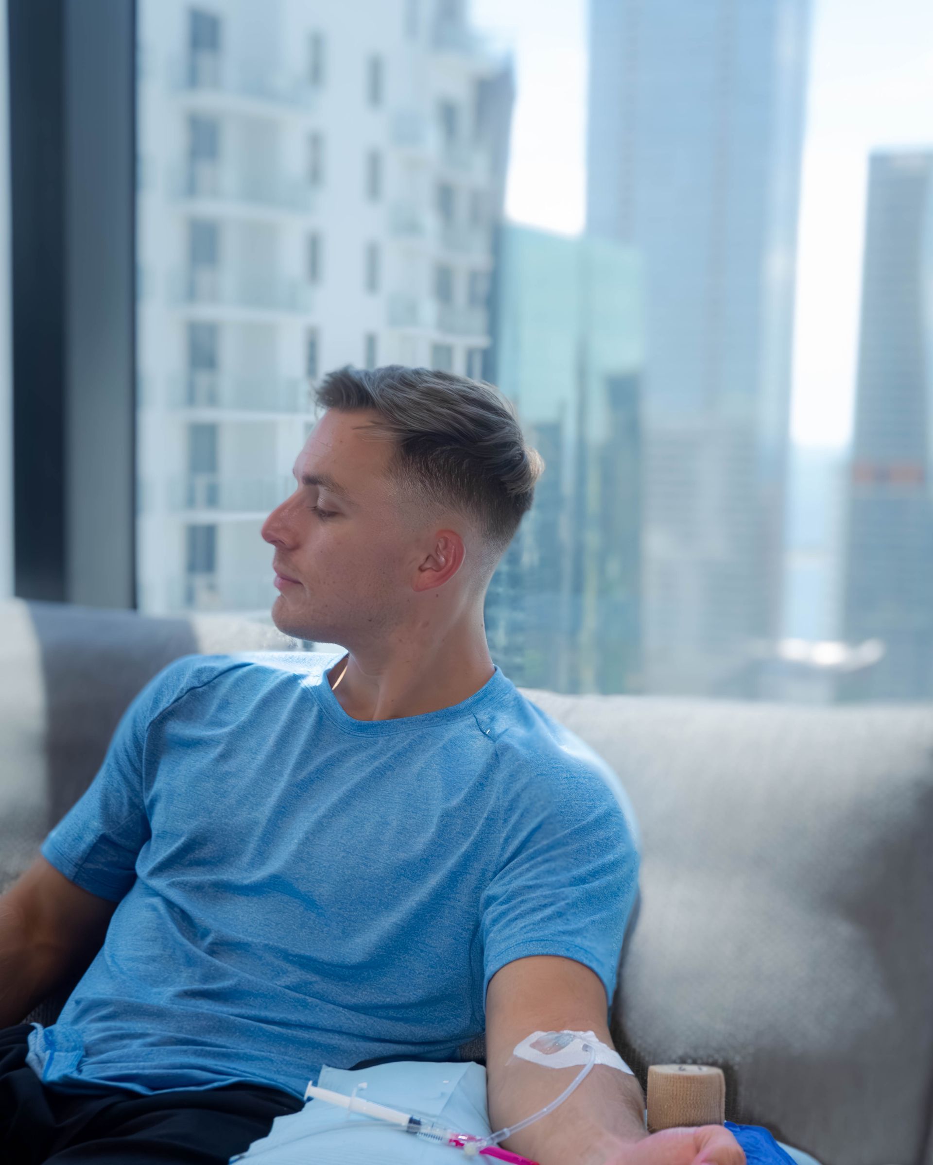 Man with IV in arm, sitting on a couch, looking out a window with a city view.
