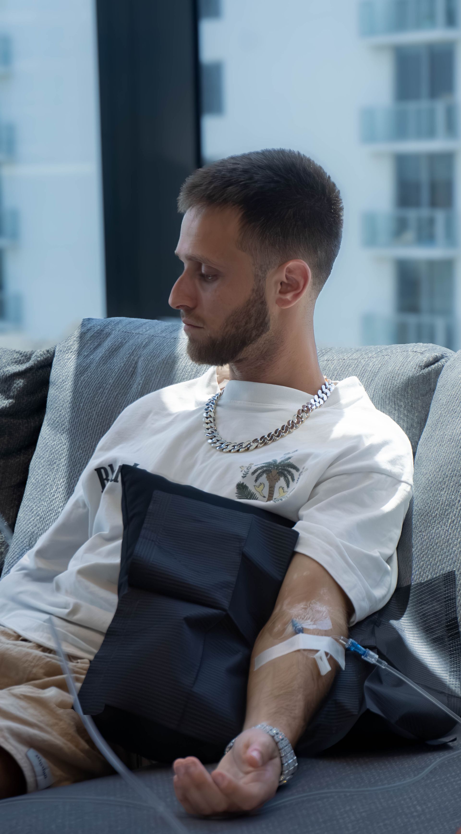 Man receiving IV drip, sitting on a couch. He is wearing a white shirt and necklace, looking down.
