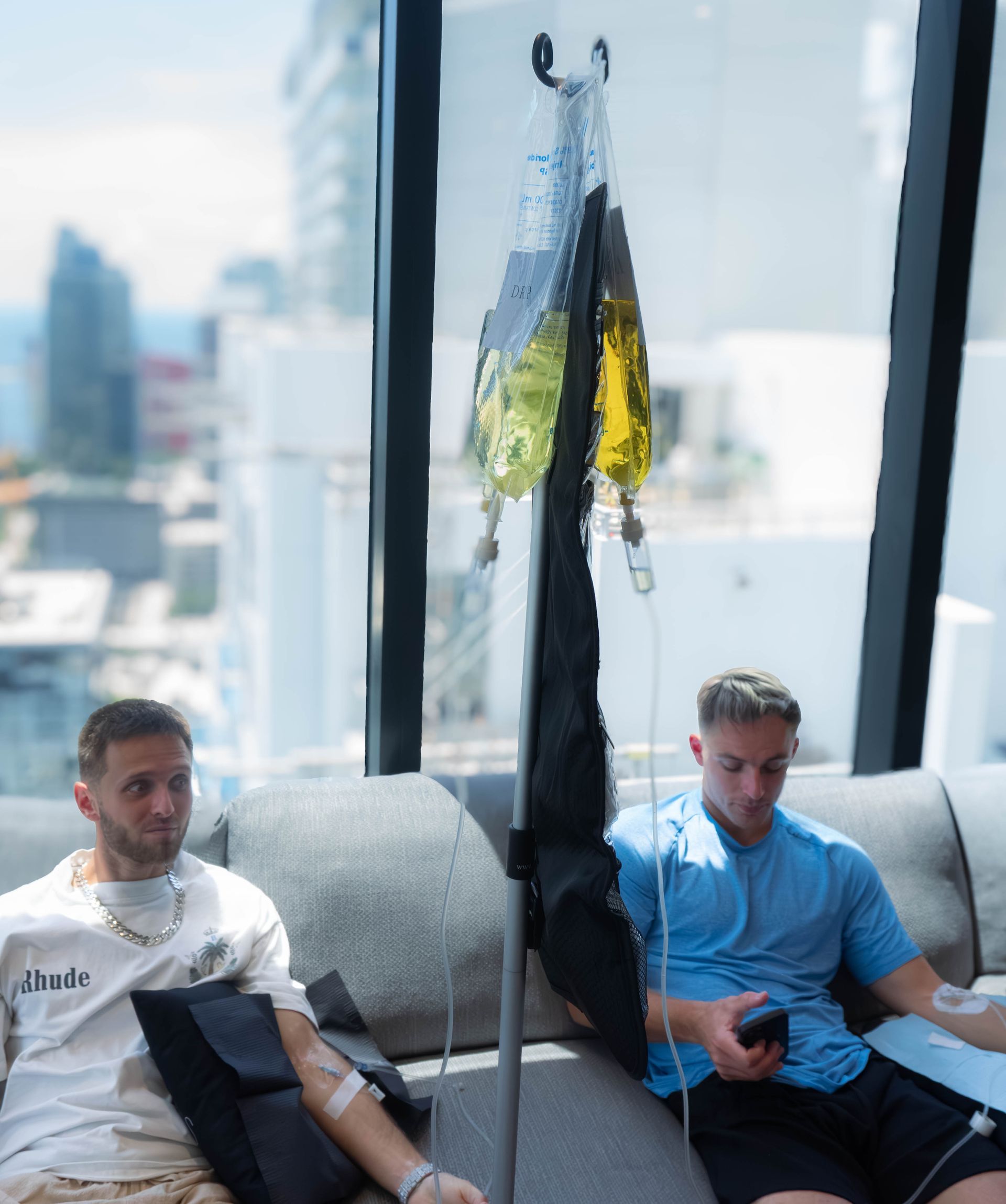 Two men receiving IV drips in a room with a city view. One man wears a white shirt, the other blue.