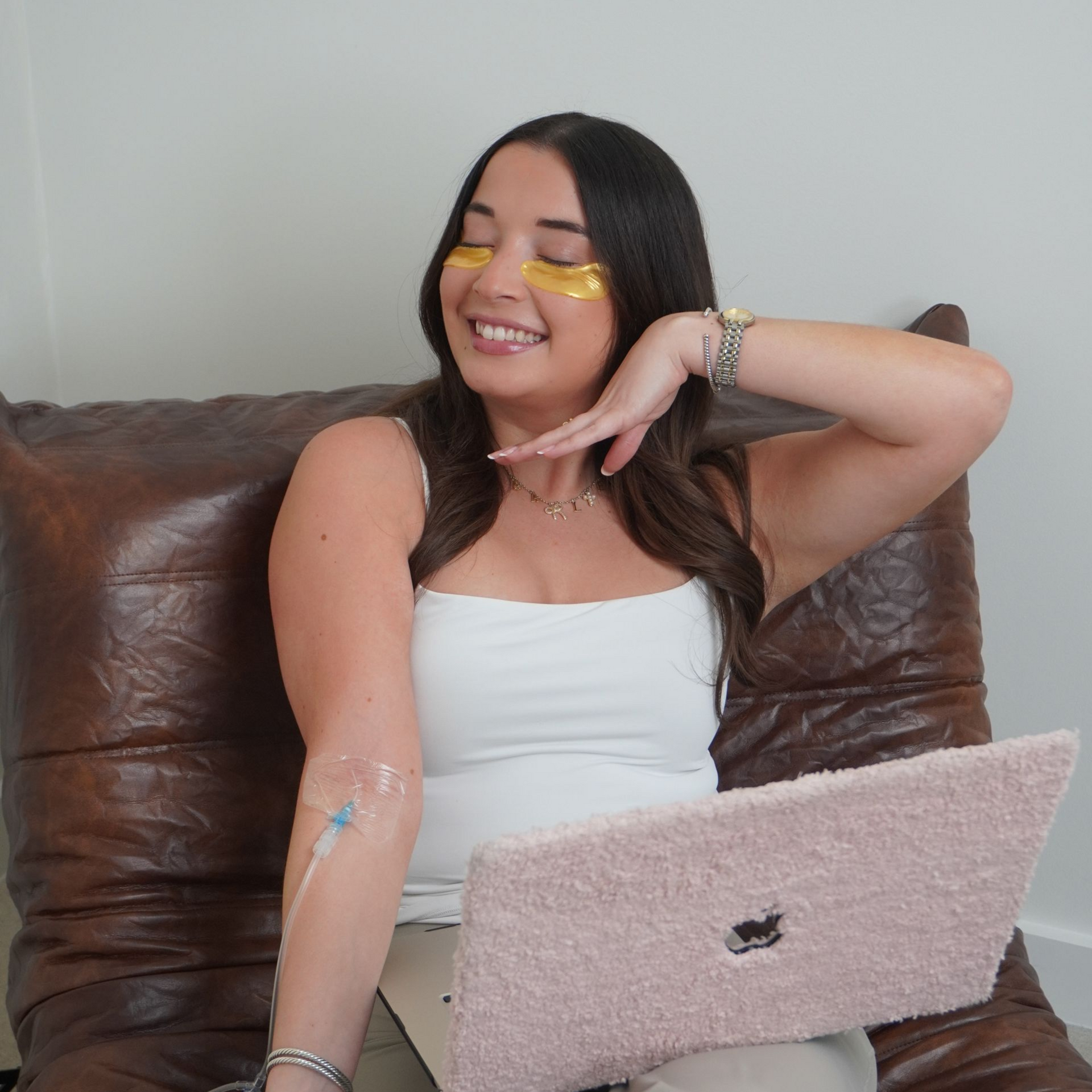 A smiling person wearing under-eye patches, posing with an IV catheter in their arm while working on a laptop.