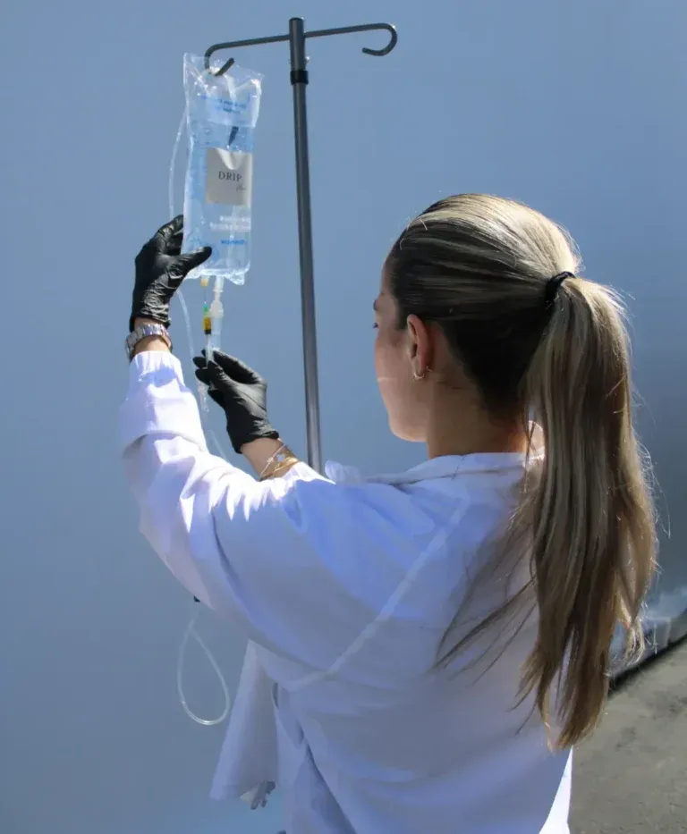 Person in lab coat prepares IV bag on a stand, wearing black gloves.