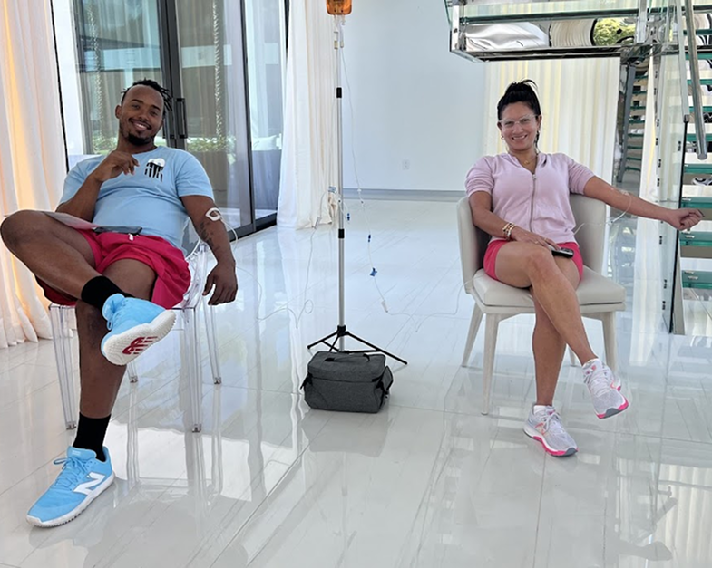 Three smiling women: one in a chair receiving an IV, two medical professionals in scrubs. Indoor setting.