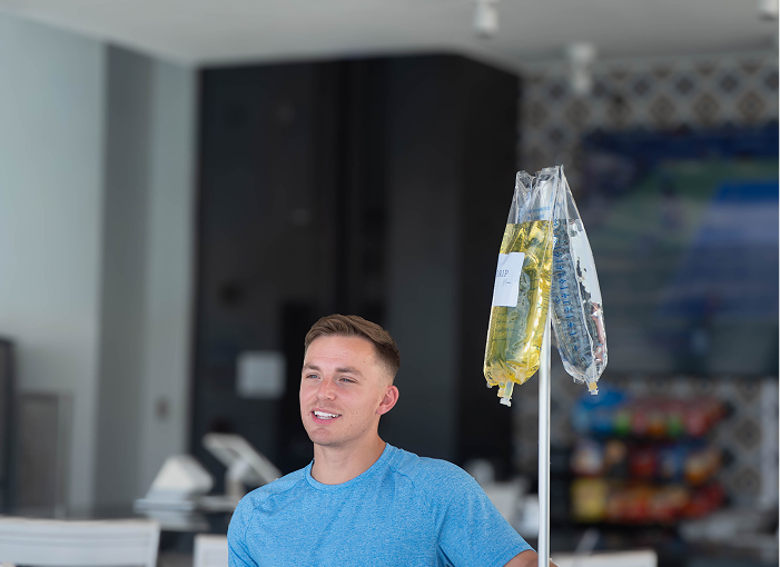 Man in blue shirt smiles, standing near IV drip in a bright room.