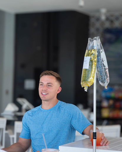Man smiling at a table with an IV bag stand. He has a drink with a straw. Bright, open interior.