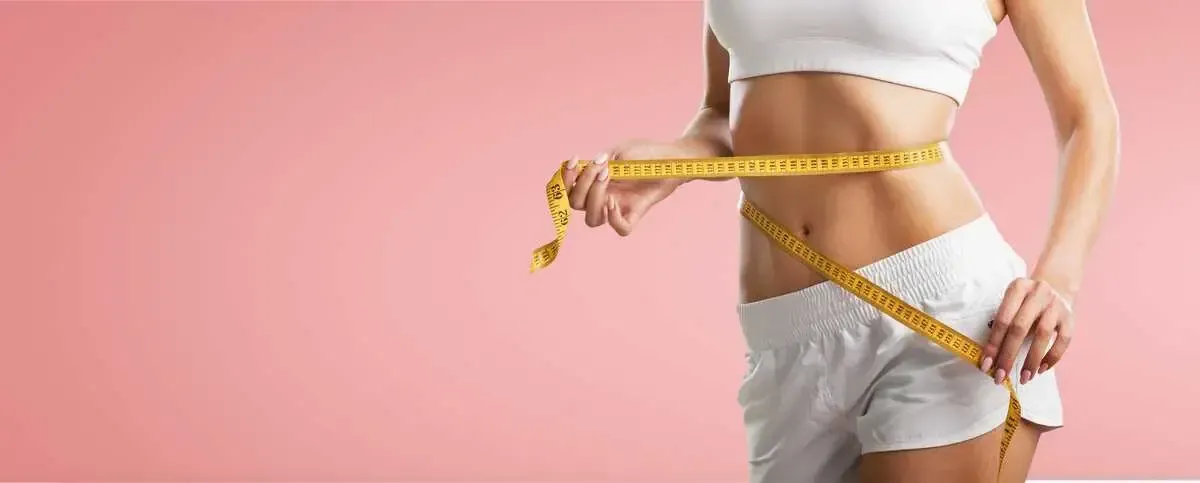 Woman measuring waist with yellow tape, pink background.