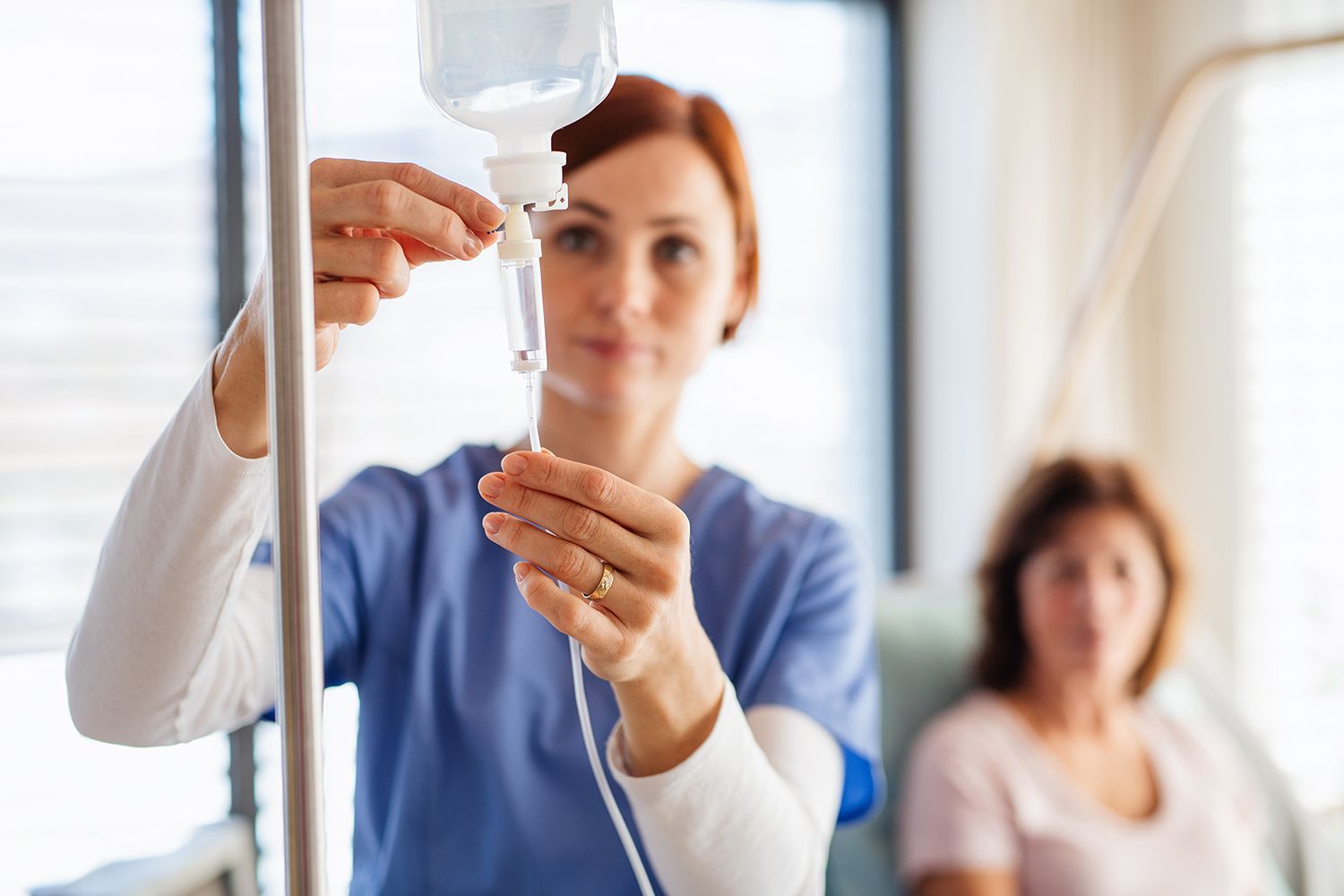 A nurse in blue scrubs adjusts an IV drip bag in a clinical setting with a patient sitting in the background.