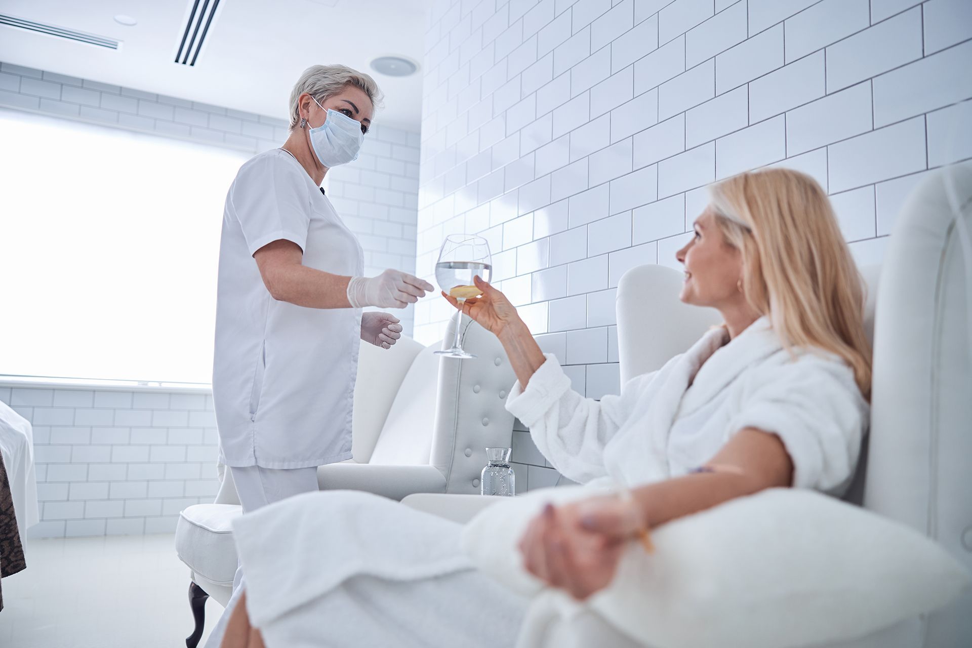 A healthcare professional in a mask and white uniform hands a glass of water to a patient in a white robe at a spa.