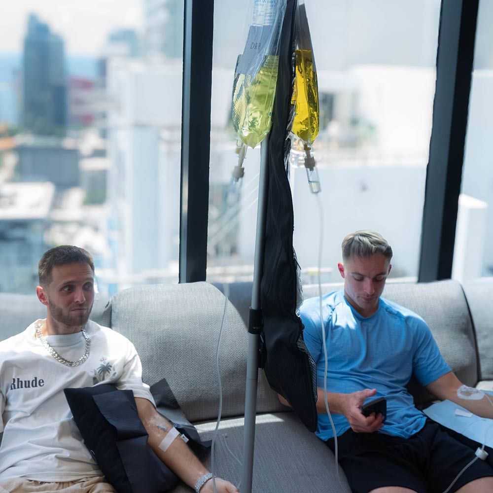 Two men receiving IV drip therapy on a comfortable couch in a high-rise setting, with a large window overlooking a sunny city skyline, for luxury wellness and recovery.