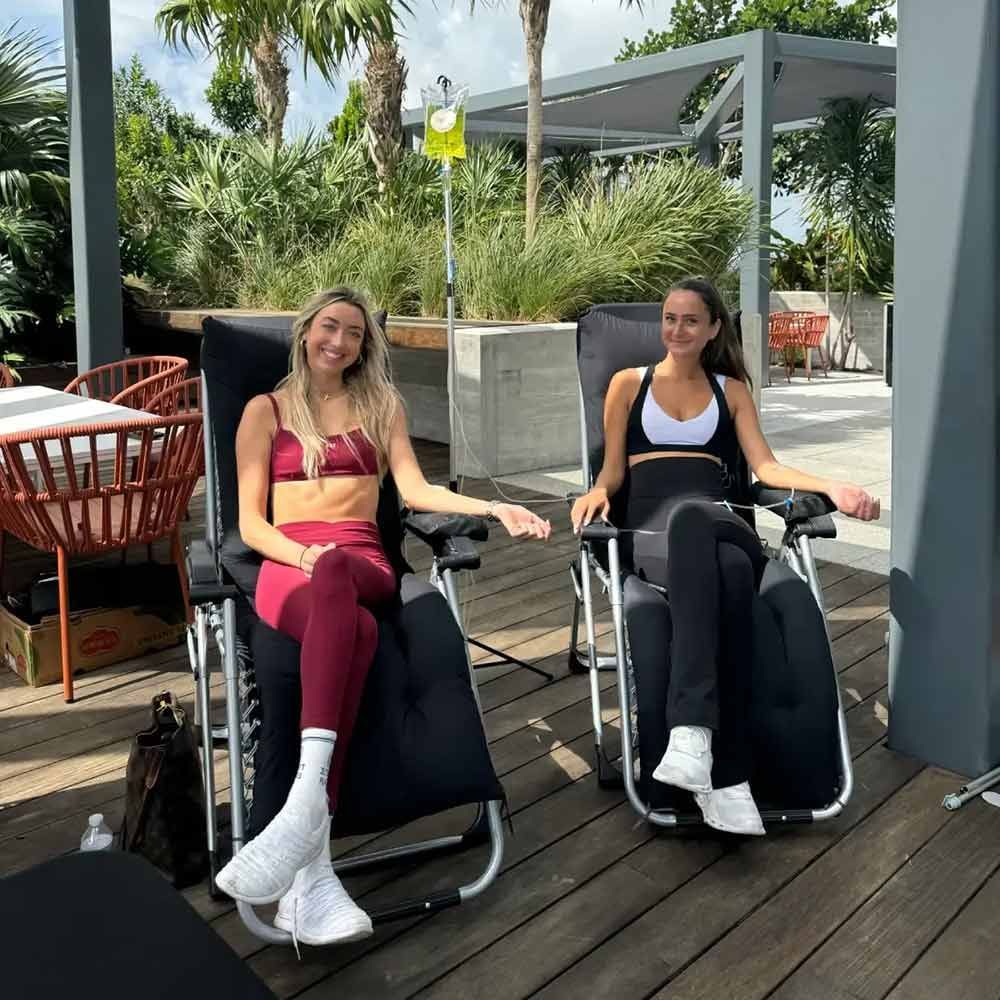 Two young women in athletic wear receiving post-workout IV recovery drip therapy in comfortable zero-gravity chairs on a sunny outdoor patio.