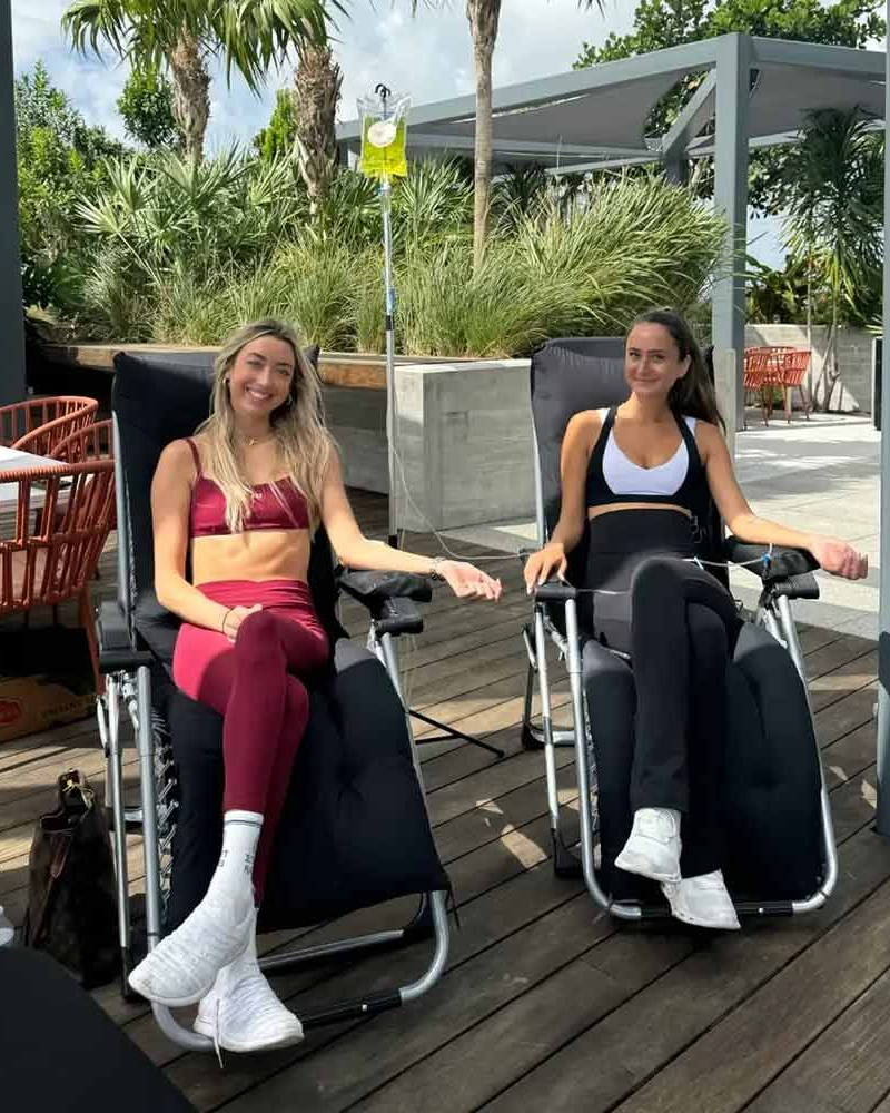 Two women relax outdoors in lounge chairs, receiving IV drips.