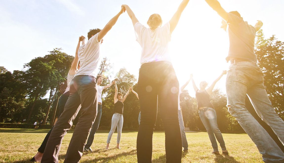People standing in a circle outdoors, holding hands with arms raised, sunny background.