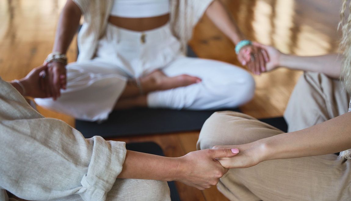 People in a circle holding hands, one person in lotus position. Interior, soft lighting, neutral colors.