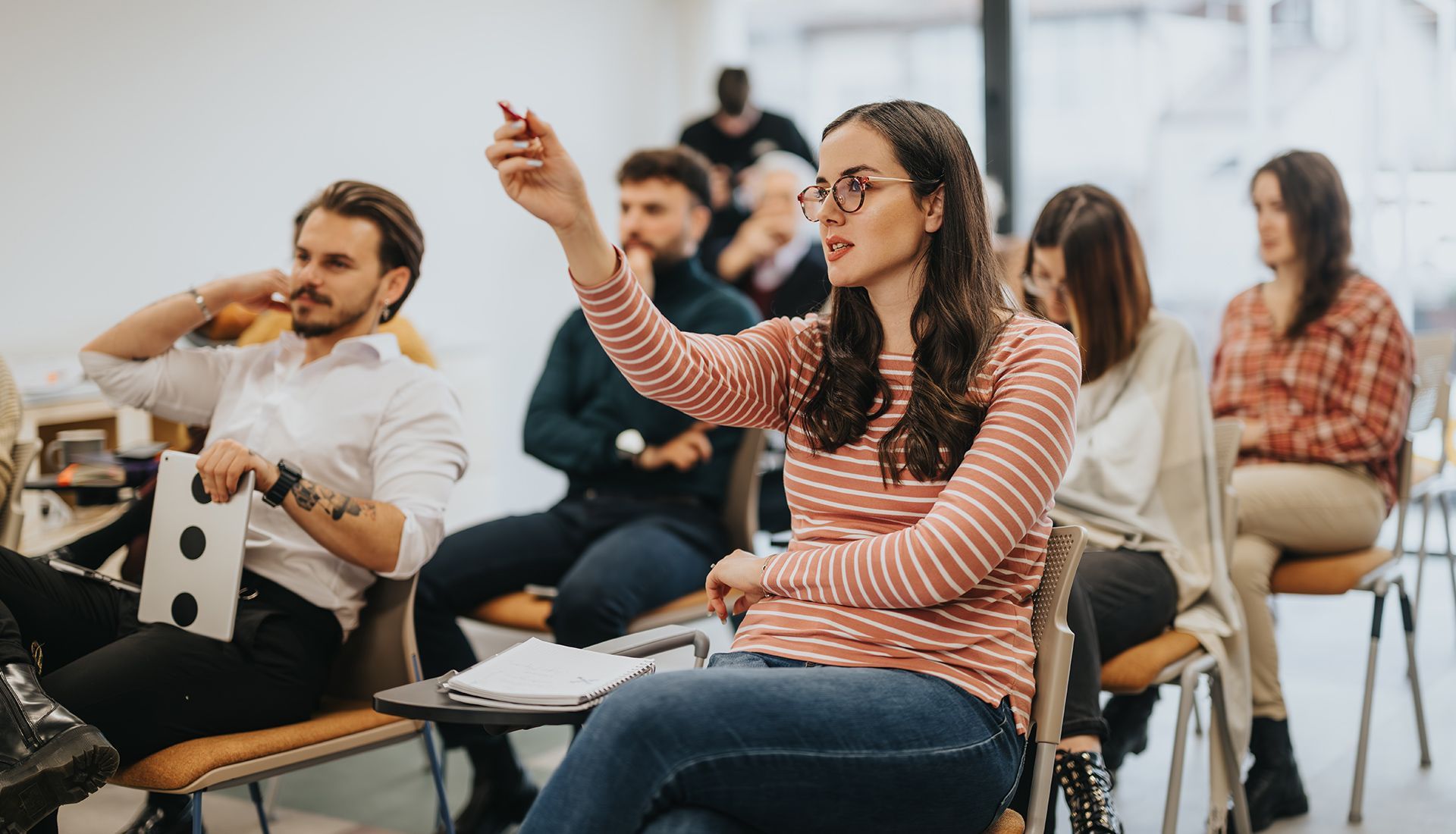 Woman in a striped shirt raises her hand, speaking in a classroom.