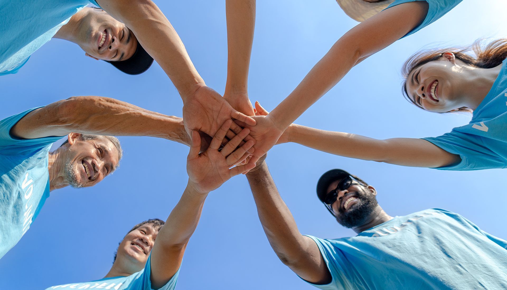 People in blue shirts with hands joined together against a blue sky, symbolizing teamwork and collaboration.