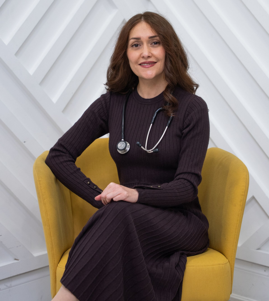 Woman seated in a yellow chair, wearing a dark sweater and stethoscope against a white geometric wall.