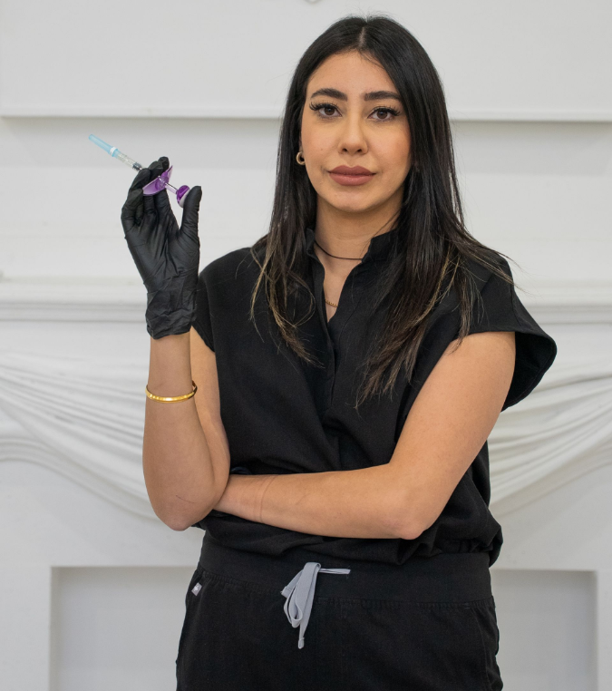 Woman in black gloves holding a syringe, standing in front of a white wall and fireplace.