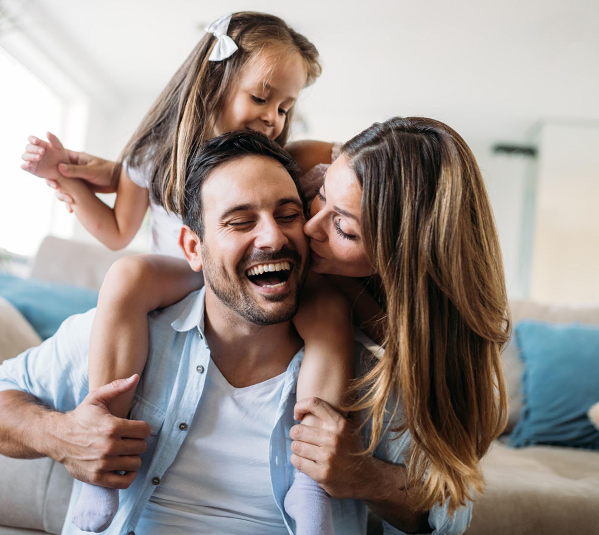 Happy family: father laughing with daughter on his shoulders while mother kisses his cheek in a living room.
