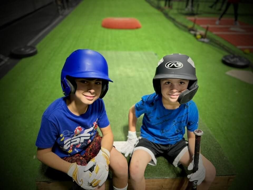 Two boys in baseball helmets and gloves sit on a bench inside, smiling.