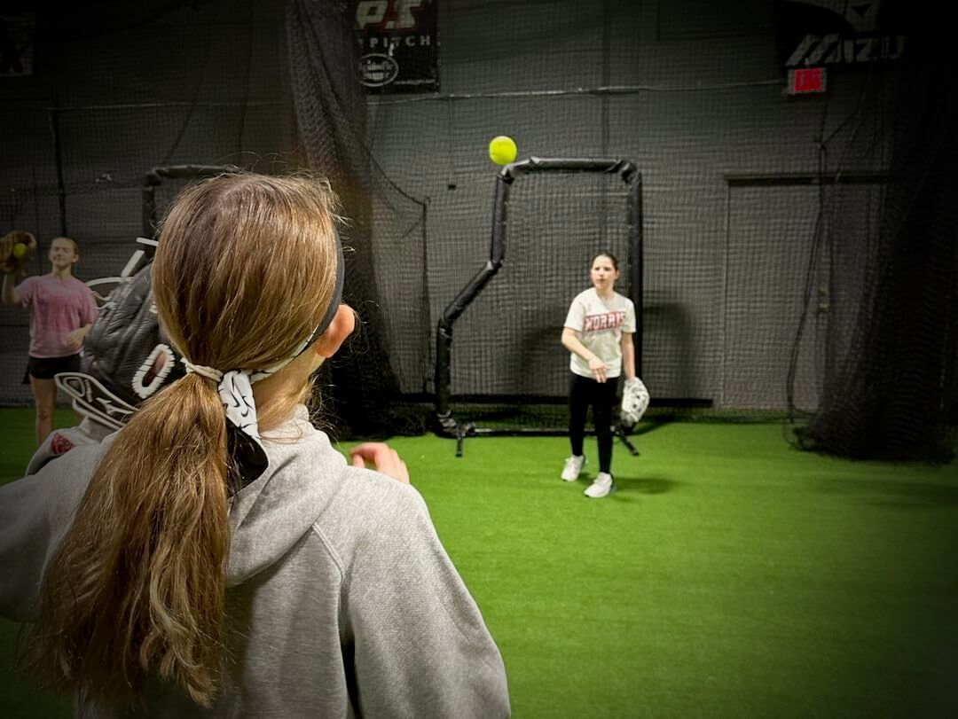 Girl in a hoodie watches a softball being pitched in an indoor practice facility; two other girls watch.