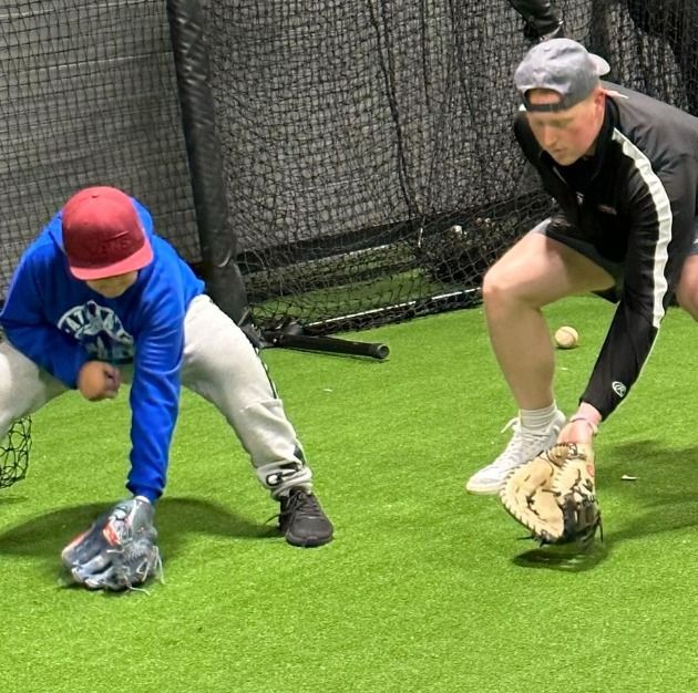 Two boys in baseball helmets and gloves sit on a bench inside, smiling.