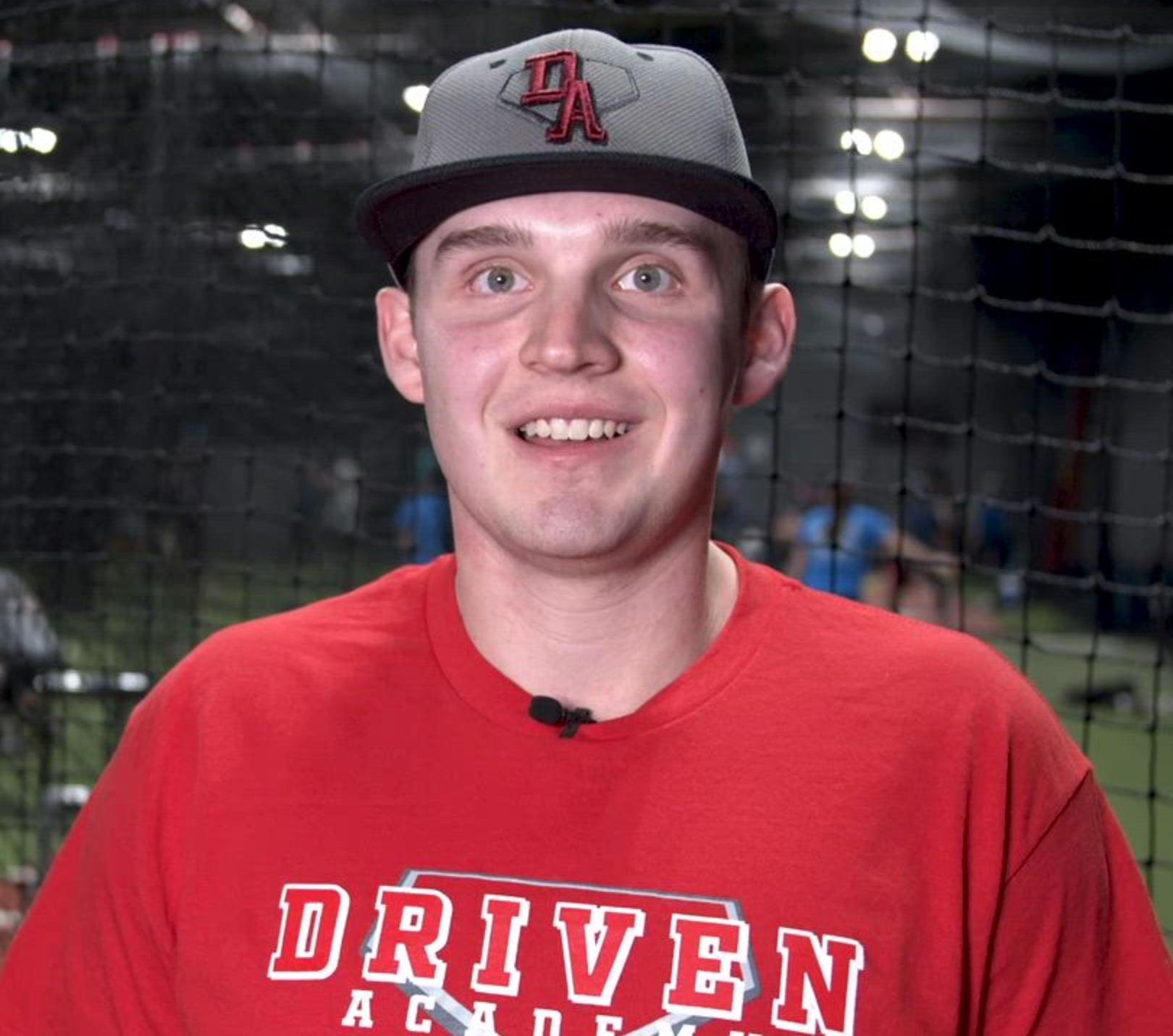 Man in red shirt and grey hat, smiling at the camera, in a baseball training setting.