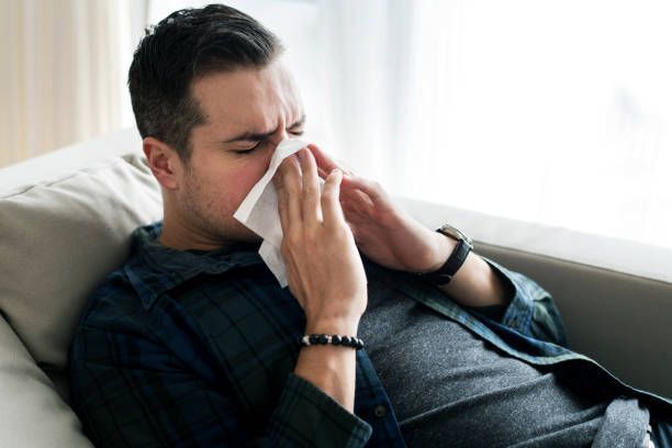 A man is laying on a couch blowing his nose into a napkin.