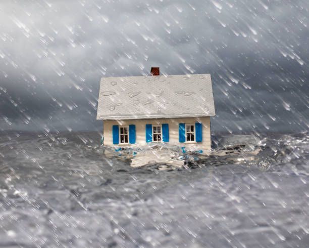 A small house with blue shutters in a stormy sea, submerged by rising water. Rain is falling.