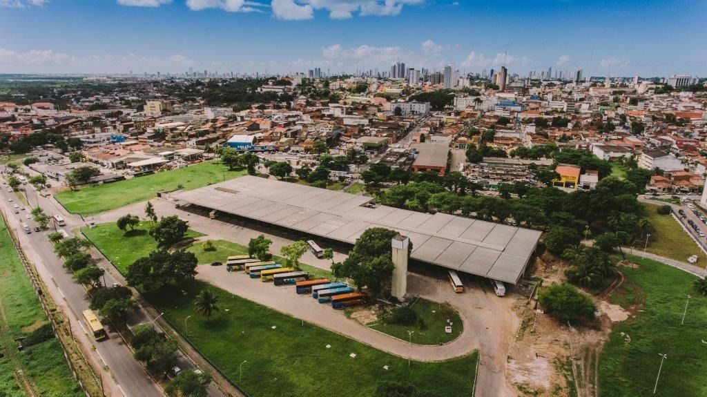 Vista aérea de uma cidade com um  terminal de ônibus e expansão urbana ao fundo, sob um céu azul.
