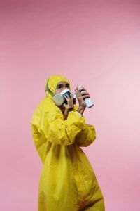 Person in a yellow hazmat suit spraying disinfectant while wearing a respirator against a pink background
