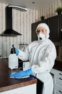 Person in white protective gear and mask disinfecting a kitchen counter with a spray bottle.