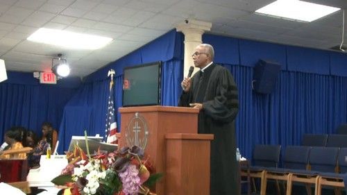 Man in black robe speaking at a podium in a church. Blue backdrop, flowers, and seating are visible.