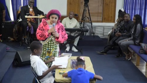 Woman in pink jacket speaks with children at a table during a church service. Musicians and audience sit nearby.