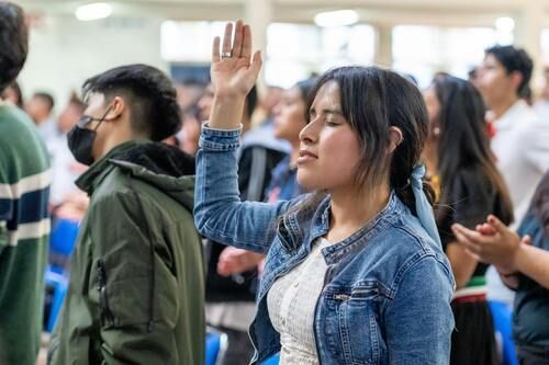 Woman in denim jacket with arm raised in worship, eyes closed. People in background.