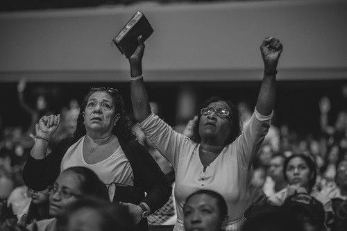 Two women in a crowd raise arms, one holding a book. Others watch in a large room.