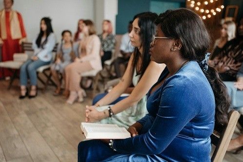 People seated indoors, listening; woman in blue dress holds book. Others in the background.