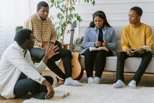 Four people sitting, holding books, in discussion; one person gesturing.