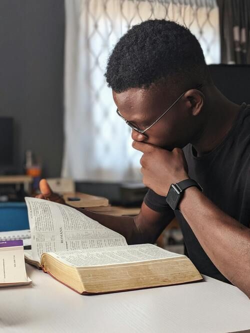 Man reading a large open book, resting chin on hand. White table, window, and blurred background.