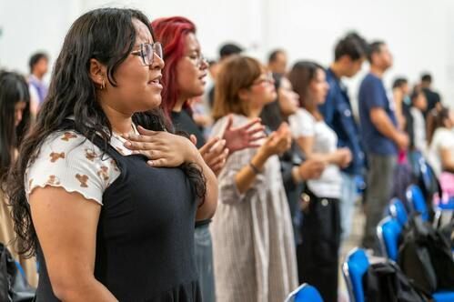 People singing with hands on chests, looking upwards in a light-filled room.