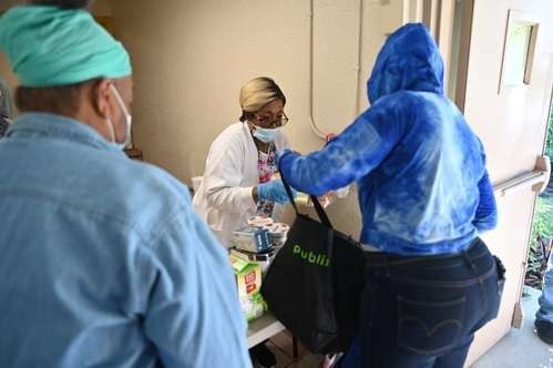 Person in blue hoodie receives a bag from a masked person behind a table, others in line.