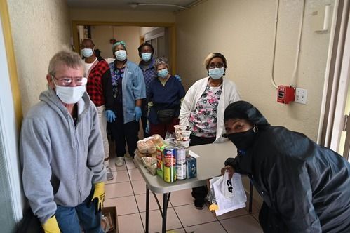 People wearing masks and gloves, preparing food packages at a table in a hallway.