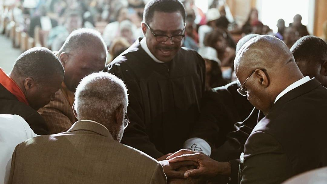 Men praying together indoors, hands clasped, solemn expressions. Sunlight illuminates the scene.