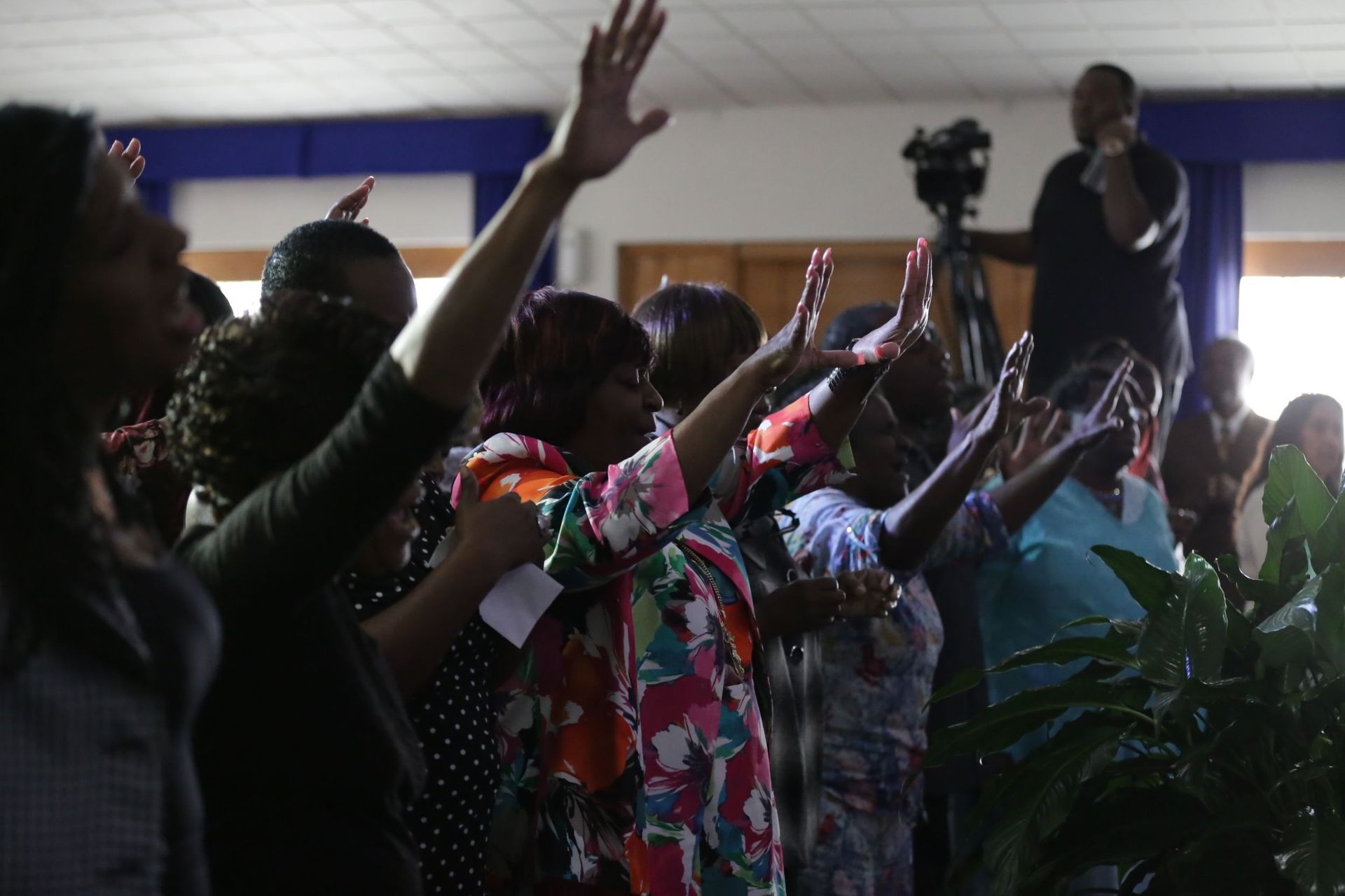 People raising hands in a room, a man preaching with camera in background.