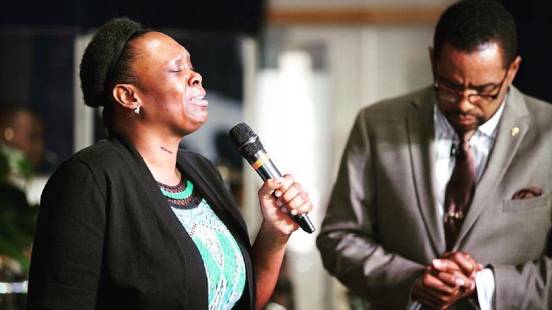 Woman singing into a microphone, man praying next to her.