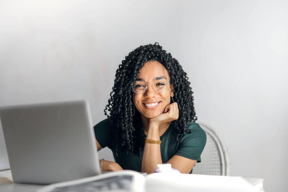 Woman with curly hair smiles while sitting at a table with a laptop and book.