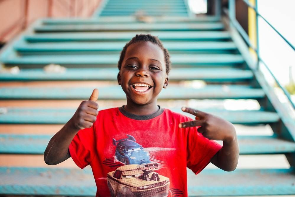 Boy in red shirt smiles, gives thumbs-up and peace sign, by turquoise stairs.