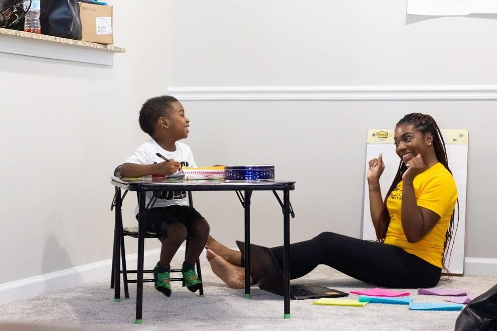 Child seated at table drawing, woman smiling and sitting on floor, learning together.