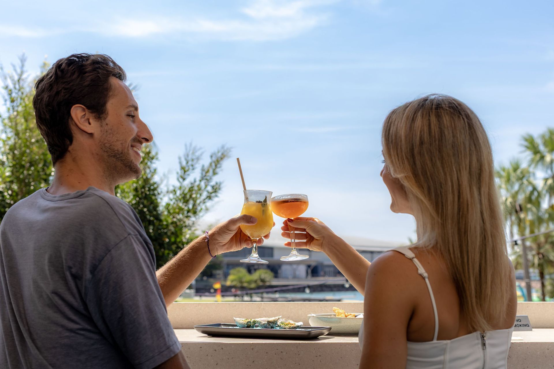 Couple clinking cocktail glasses, smiling, outdoors. Sunny sky, trees, and small food tray.