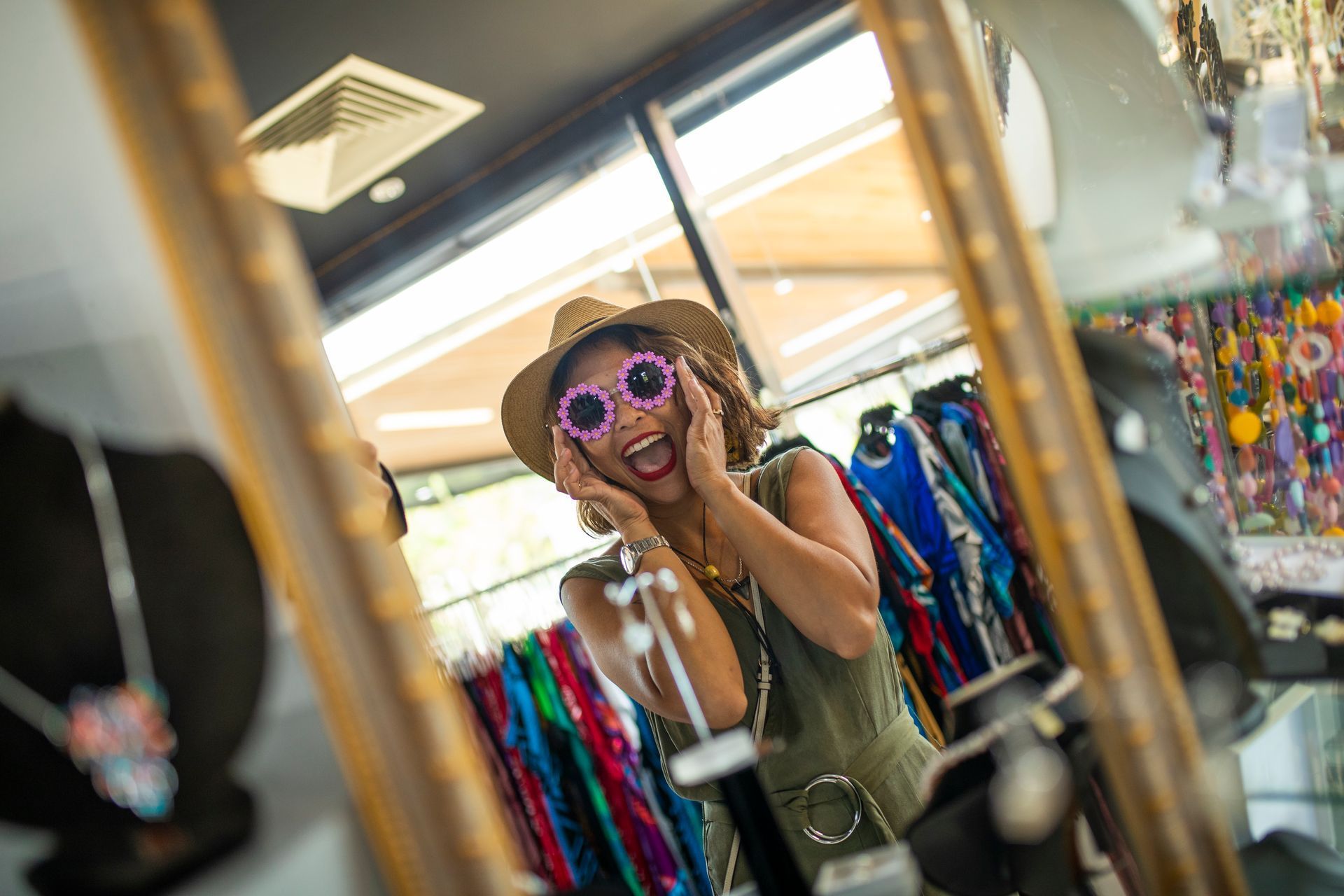 A woman faces a mirror with her hands either side of her face, she is smiling and wearing funky pink sunglasses. 