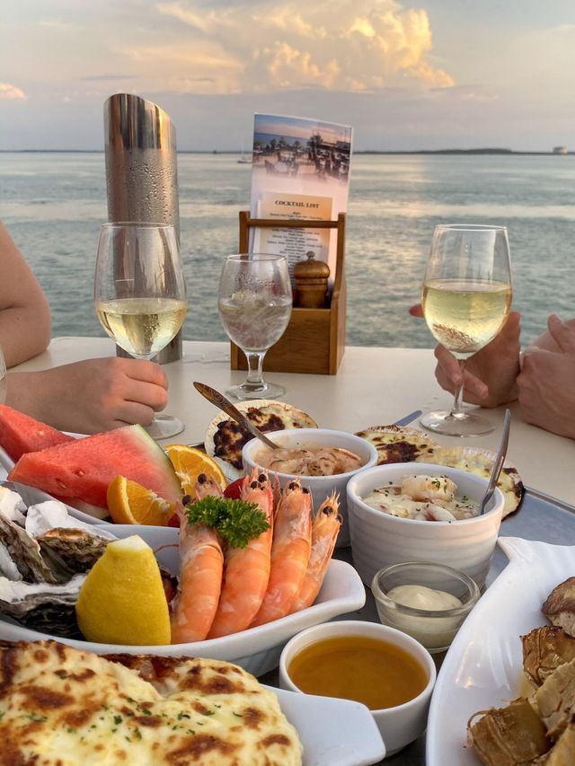 Seafood spread on a table, with wine glasses, overlooking the water at sunset.