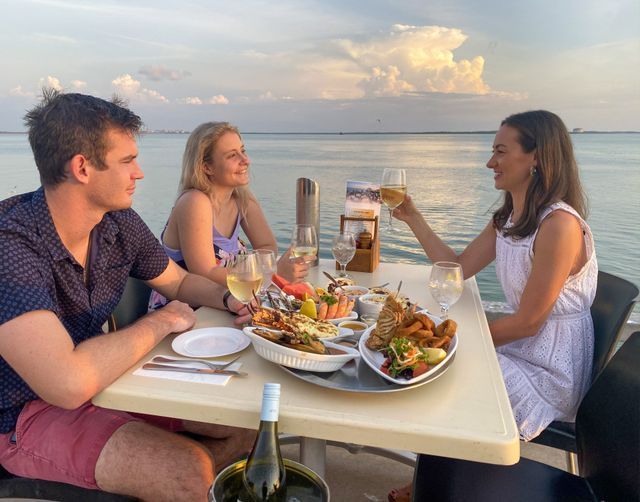 Three people dining at a waterfront restaurant, toasting with drinks; seafood platter on the table.
